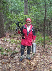 A researcher holds a crossbow and metal container of liquid nitrogen in a forest. 