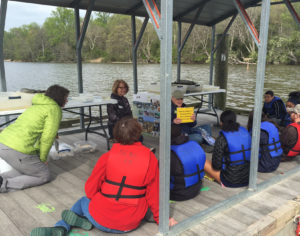 Seated man talks to kids in life jackets on floating dock.