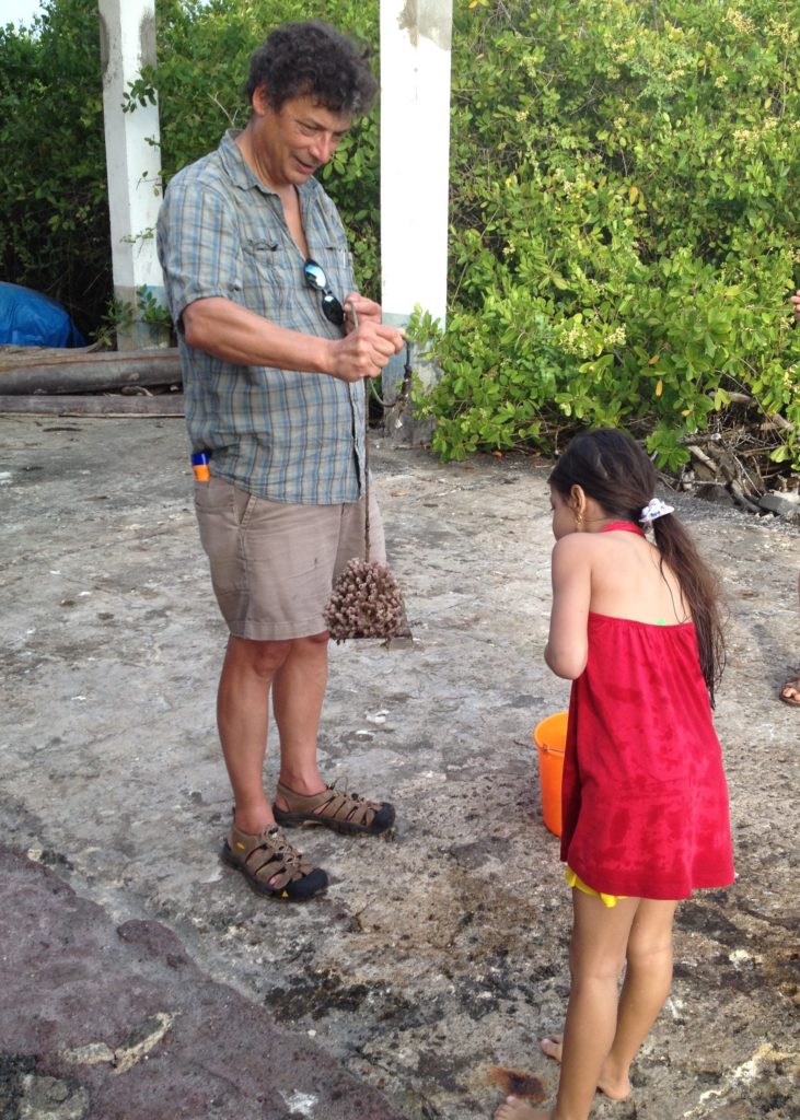 Scientist iholds a sponge attached to a rope in front of a young girl in a red dress n the Galápagos.