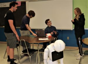 Three high school boys talk to a young woman with a white robot in the foreground.