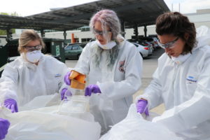 Three women in white jumpsuits sort through trash bags