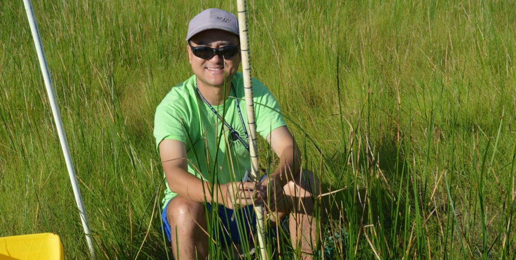 Man in sunglasses and green T-shirt sitting in a marsh holding a measuring stick