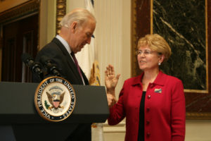 Man at lectern with presidential seal beside woman in red blazer, with her right hand raised.