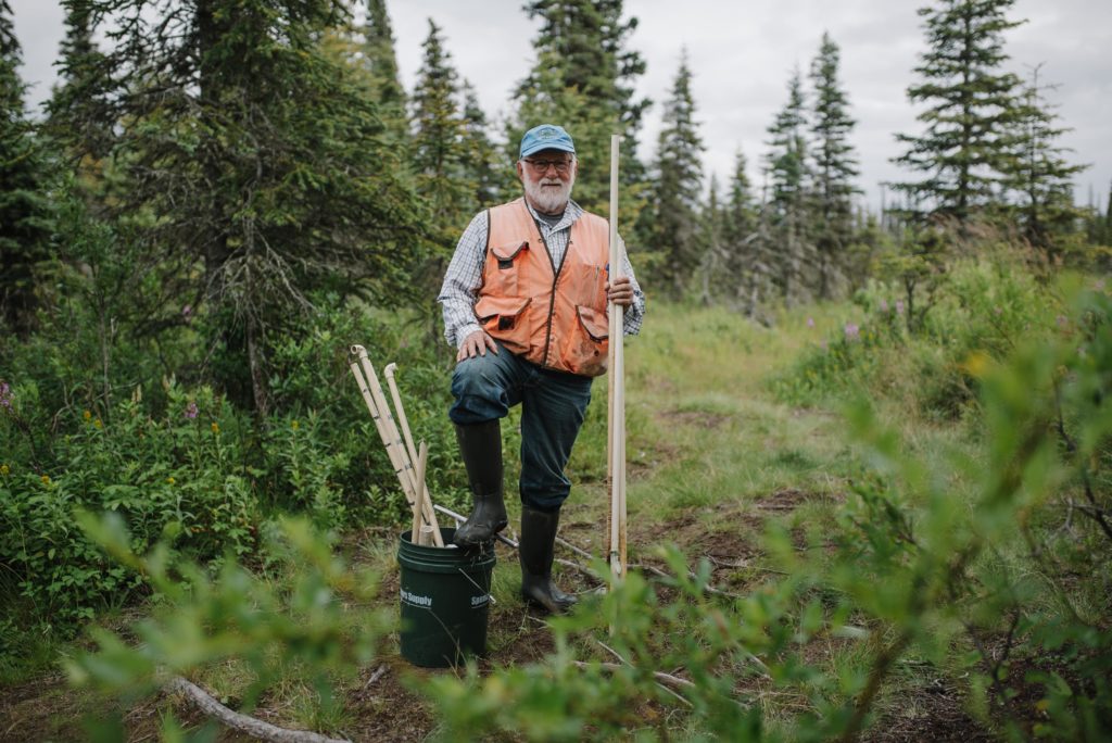 Man with a white beard and orange vest stands beside a grassy streambed holding PVC pipes.