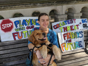 Teenage girl in blue tie-dye shirt sitting on a bench holding a golden retriever. Posters behind her read "Stop playing with our future" and "Fridays for Future" in rainbow marker.