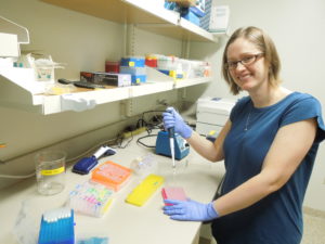 Blonde woman in blue shirt and glasses standing in lab holding pipette.