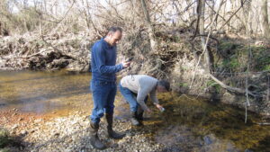 Two men in a shallow stream, one kneeling to sample water and one holding a small electronic device.