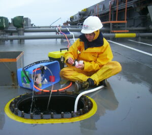 Young woman in a yellow jumpsuit and hard hat sitting on a ships deck, taking samples from an open tank