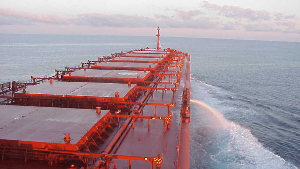 Photo taken from onboard the deck of  a massive red cargo ship. The ship juts out into the ocean, with no land in sight. A fountain of ballast water arcs out the right side of the ship.