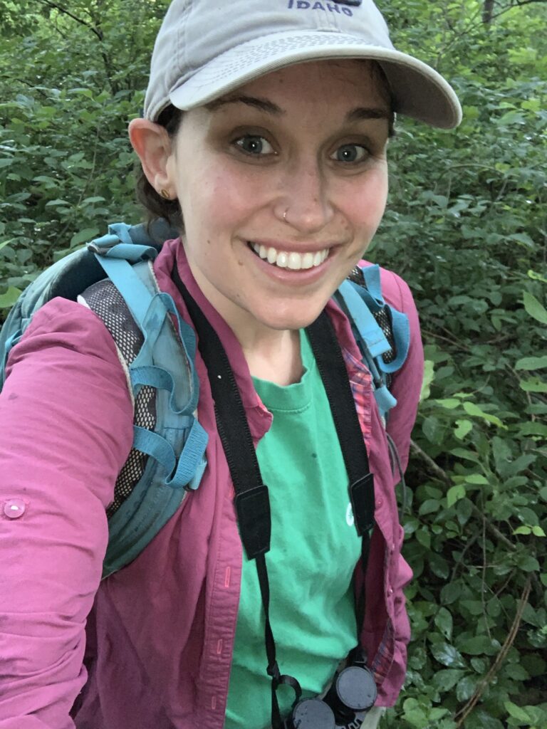 Selfie of a scientist hiking through a forest, wearing a blue backpack and a white baseball cap