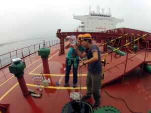Two people stand on a red ship examining a bottle of water sampled from the ballast tank.