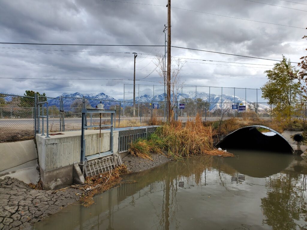 A brown urban stream flows beside a road, with snow-capped mountains in the background