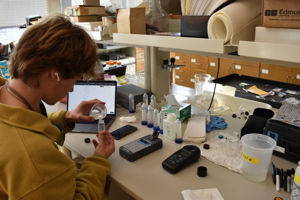 We see a laboratory bench top full of test tubes with water samples and various small machines scattered about. Parker Franco, high school volunteer, carefully pours water from a beaker into a small vial to read on the AquaFluor machine which measures chlorophyll and CDOM content. He listens to music on his airpods and has a laptop open with the data entry template in Fieldscope (the website the project uses to report data) on the screen. 