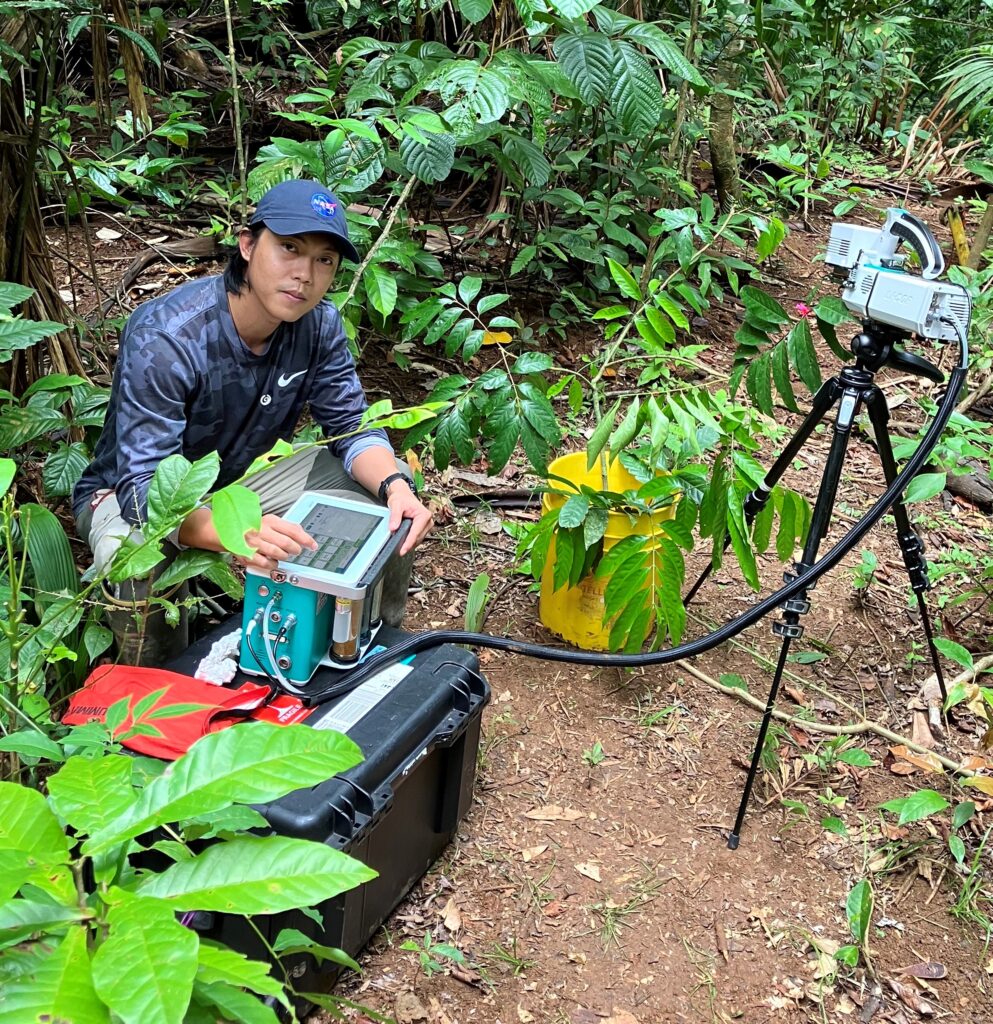 Young man kneeling in a rainforest. He is holding a small scientific measuring device on a black crate, connected to another scientific instrument on a tripod.