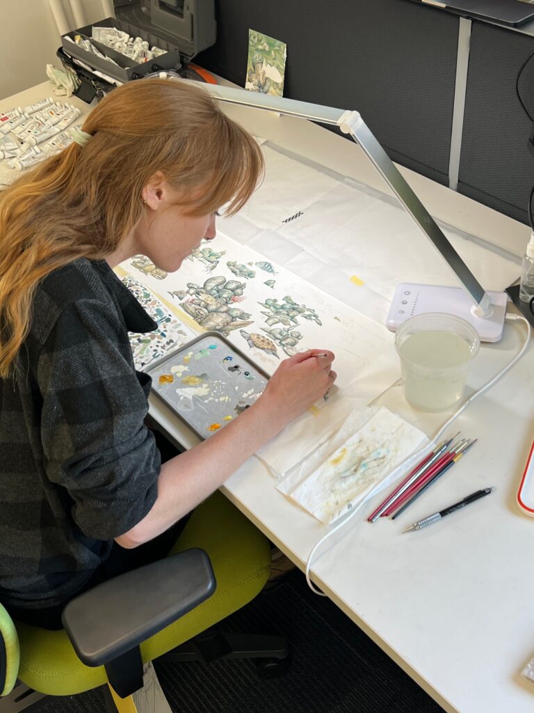 Young woman with long red hair sits at a desk with science illustration tools, painting turtles, fish and other aquatic animals