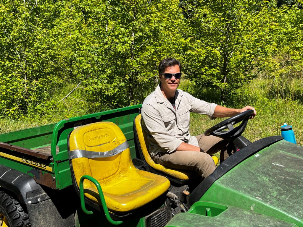 John Parker, wearing a beige shirt and sunglasses, sits in a green gator in front of a forest plot in BiodiversiTREE