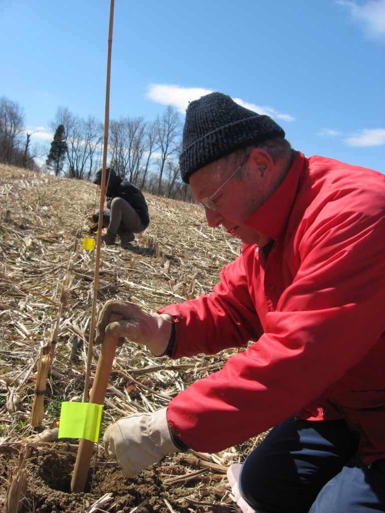 Man wearing a red winter jacket and black hat kneels in a brown field, using a wooden stick to round out a hole