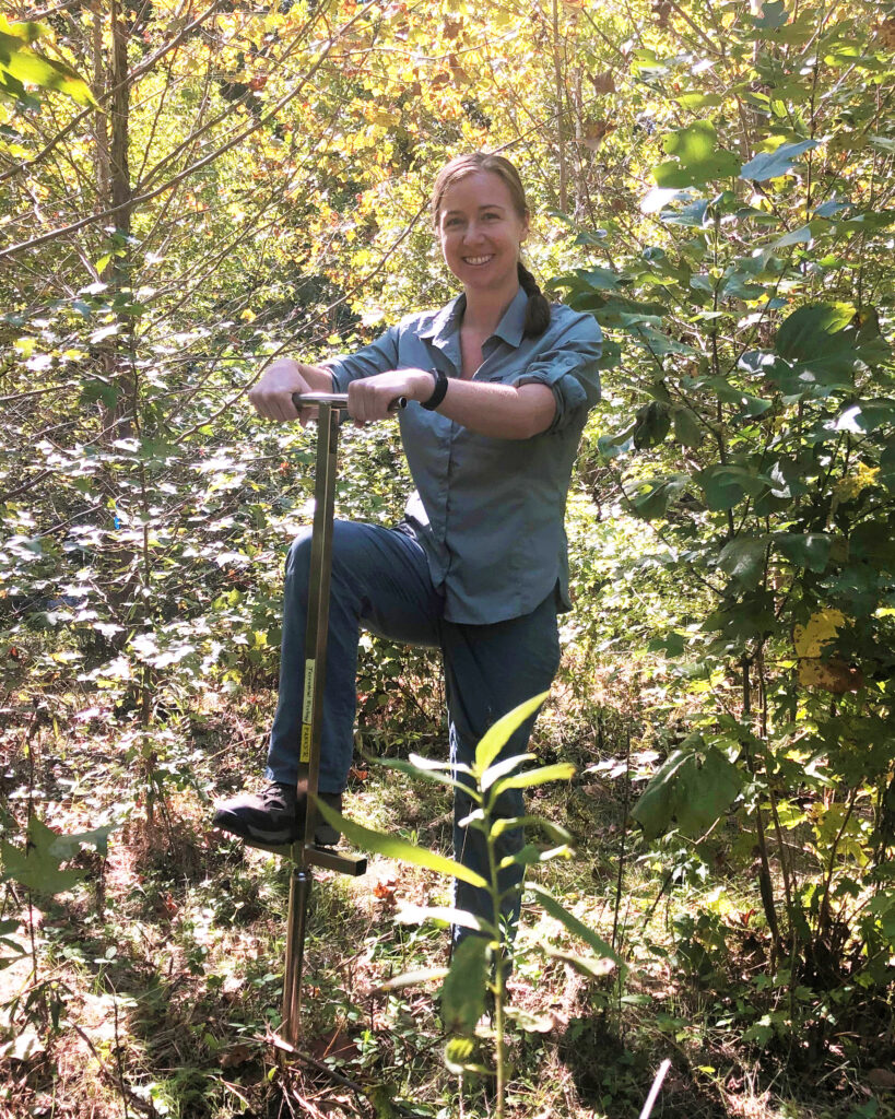 Rachel King, a young woman with long brown hair wearing gray and blue field clothes, stands with one foot on a soil corer in a patch of trees