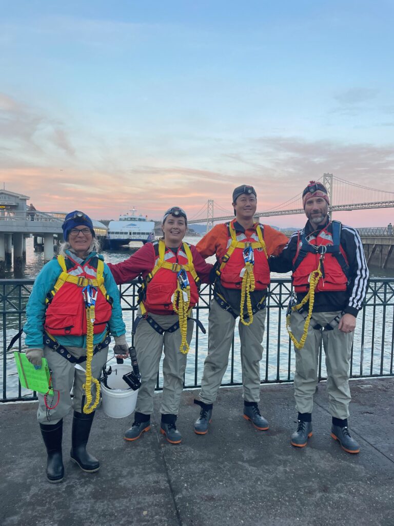 Four scientists in orange vests and safety harnesses smile with their arms around each other, with the city waterfront behind them.