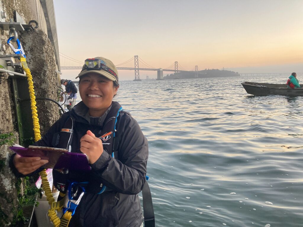 Young woman stands partially in the water beside a seawall, wearing a safety harness and holding a clipboard. She is smiling at the camera, with the Golden Gate Bridge in the background