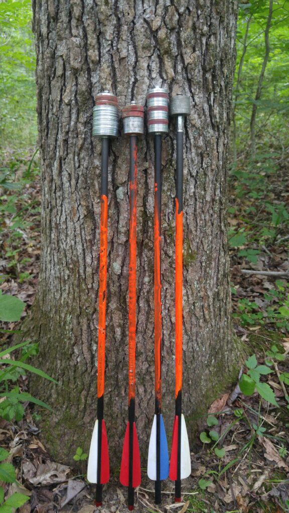 Four orange crossbow bolts rest against a tree, their tips covered with stacks of silver and red washers.