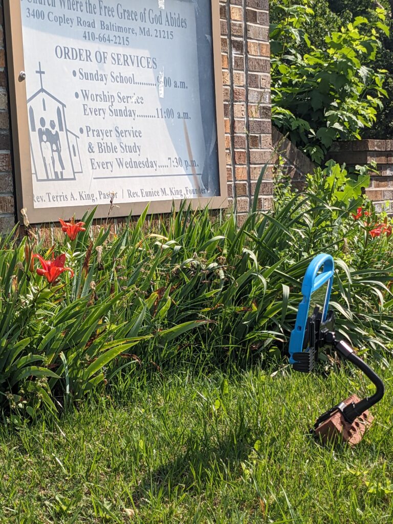 A blue iPad sits on the grass, propped up on a stand with a curved neck. It faces a church sign with service times and the words "Church Where the Free Grace of God Abides"