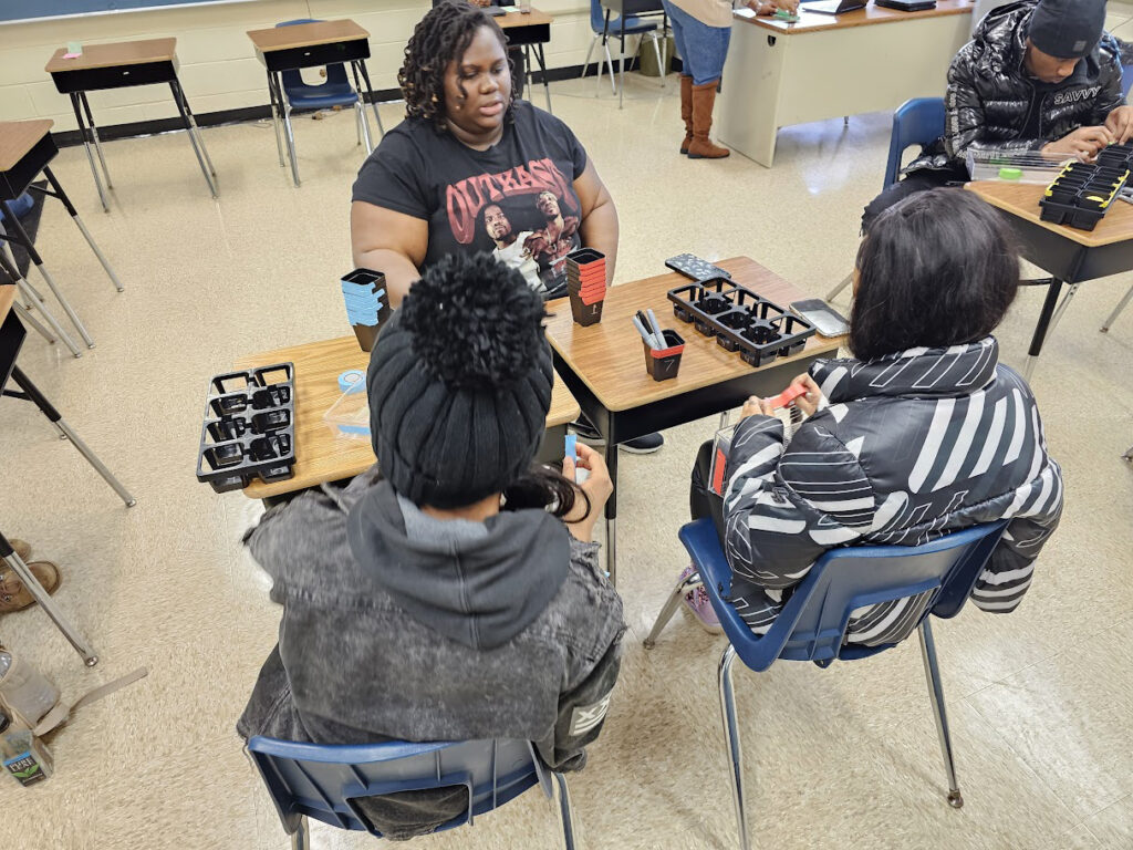 Shatiyana Dunn, a young woman with dark brown skin, sits across from two Classroom Cultivation students at their desks with empty black trays for plants.