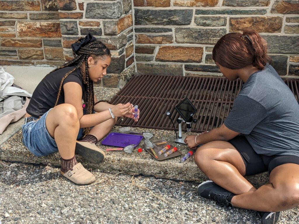 Two teenage girls with light brown skin sit next to a sewer grate beside a multicolored brick brick wall. Clear plastic vials and other scientific equipment are on the ground between them.