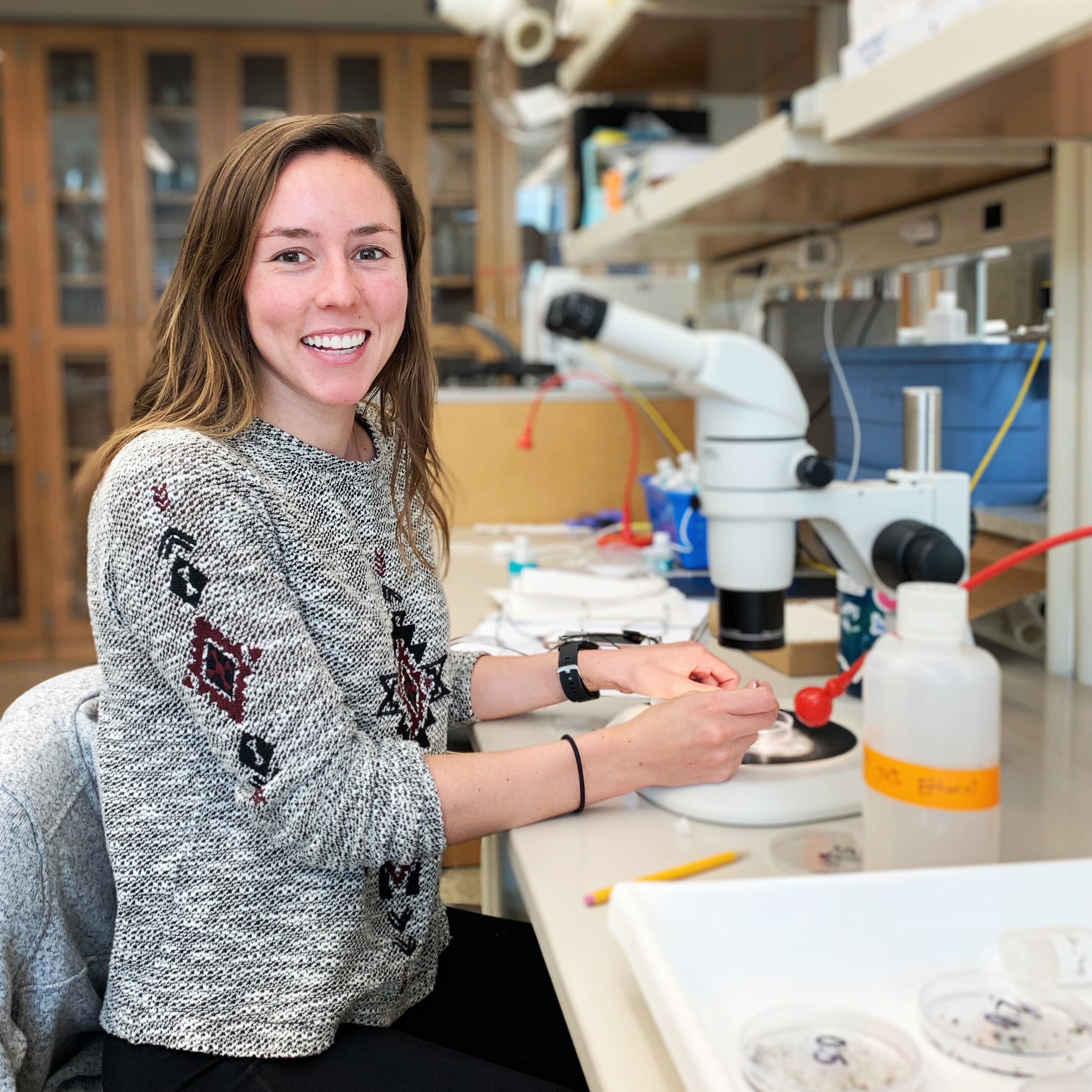 A young woman wearing a gray sweater sits at a lab table, smiling at the camera while holding samples under a microscope.