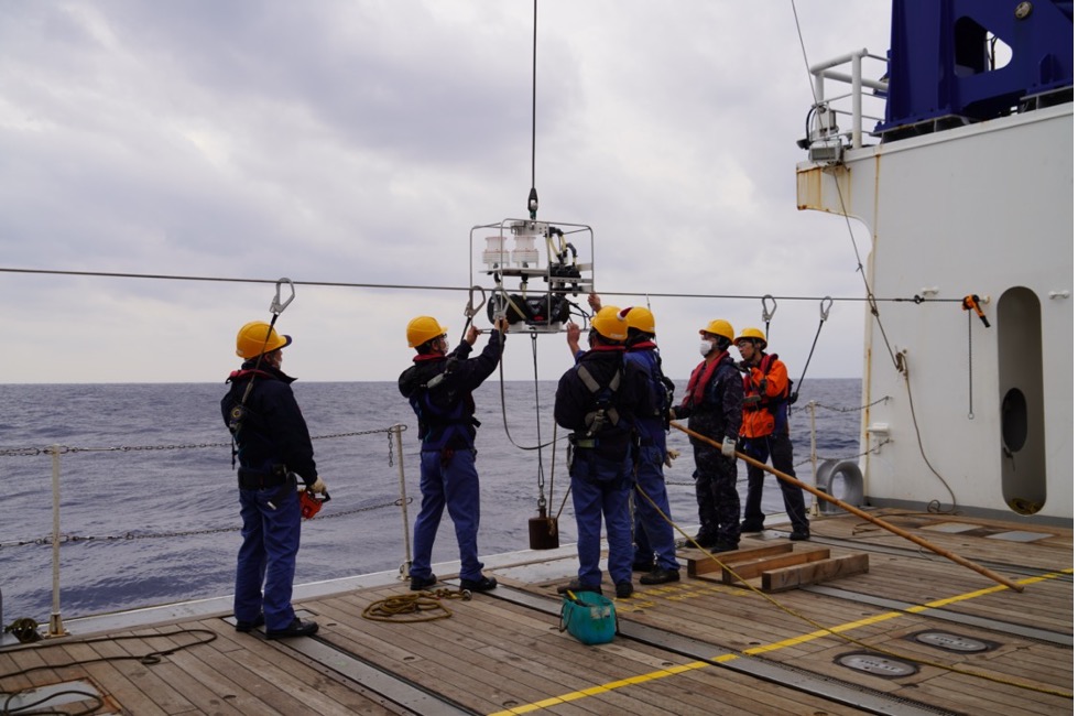Six people wearing yellow hard hats stand on the deck of a boat, preparing to deploy a scientific instrument suspended in the air.