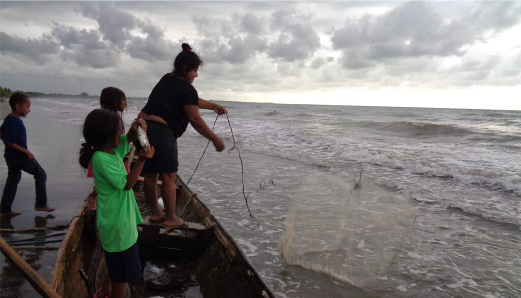 A woman and two children stand on a boat in shallow water while another child walks behind the boat. The woman holds a line attached to a fishing net over the boat, while the child in front holds a silver fish.